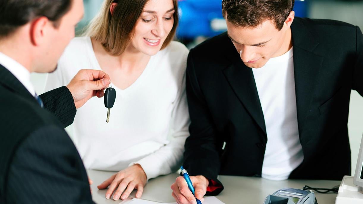 A man and woman signing financing documents while a salesperson hands them a set of keys